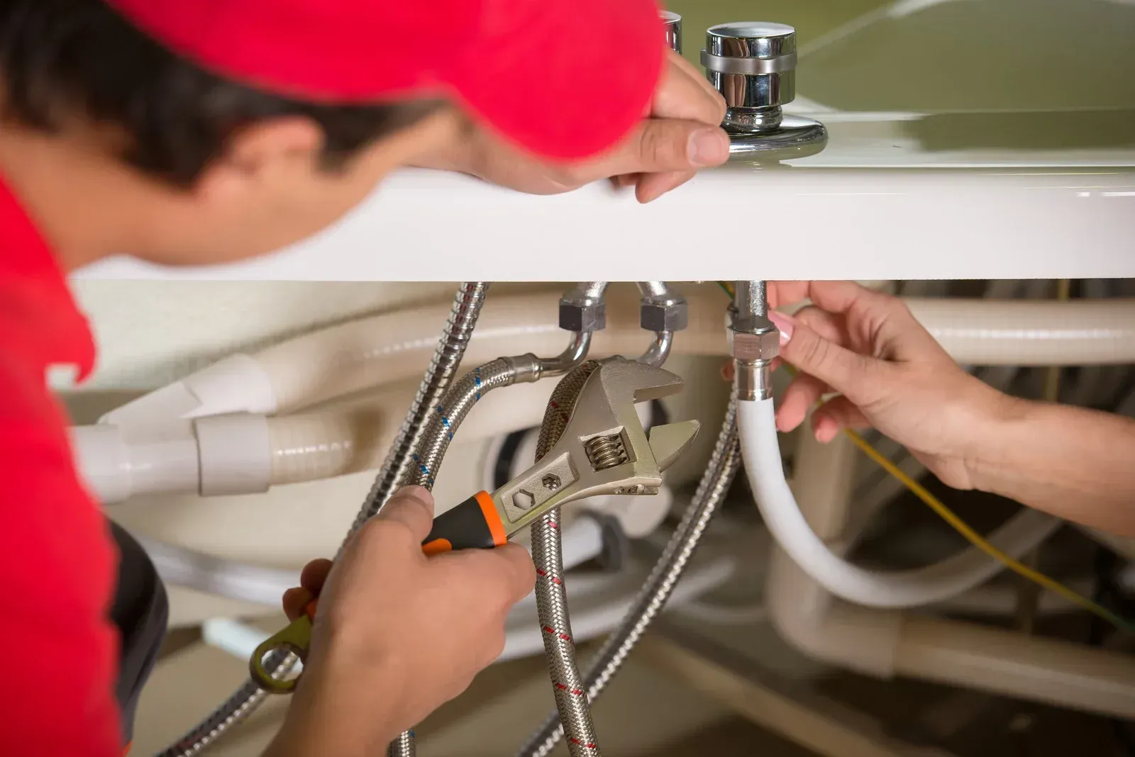 Plumber in red shirt and hat uses wrench to fix pipes under a sink.