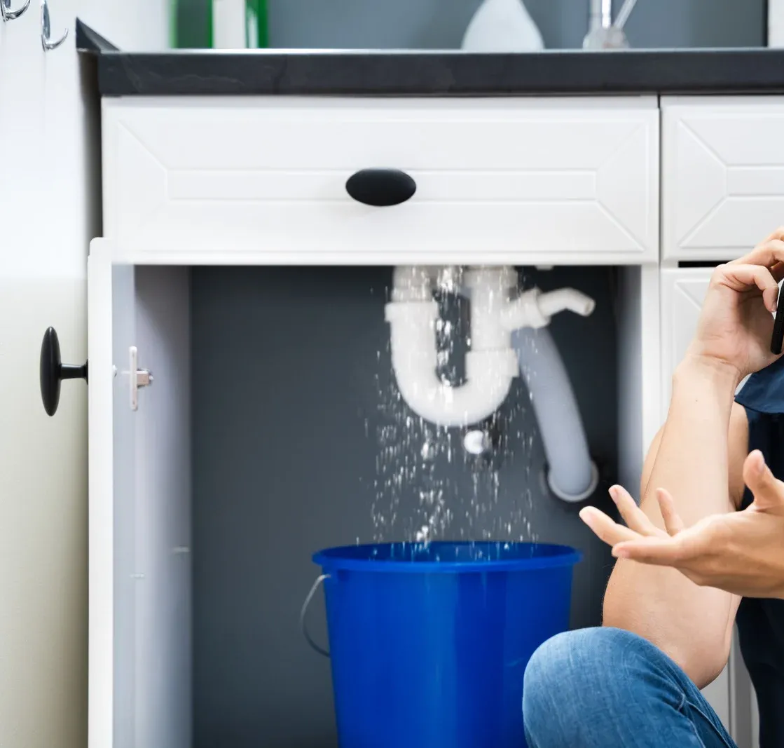 Man reacts to water leaking from a sink's pipes into a bucket inside a cabinet.