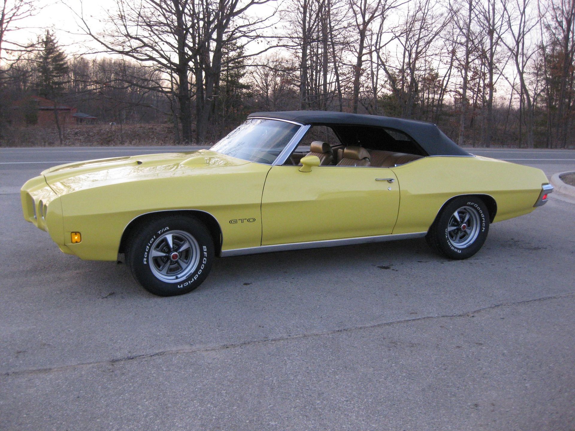 Yellow 1970 Pontiac GTO convertible with a black top on a paved road with trees in the background.