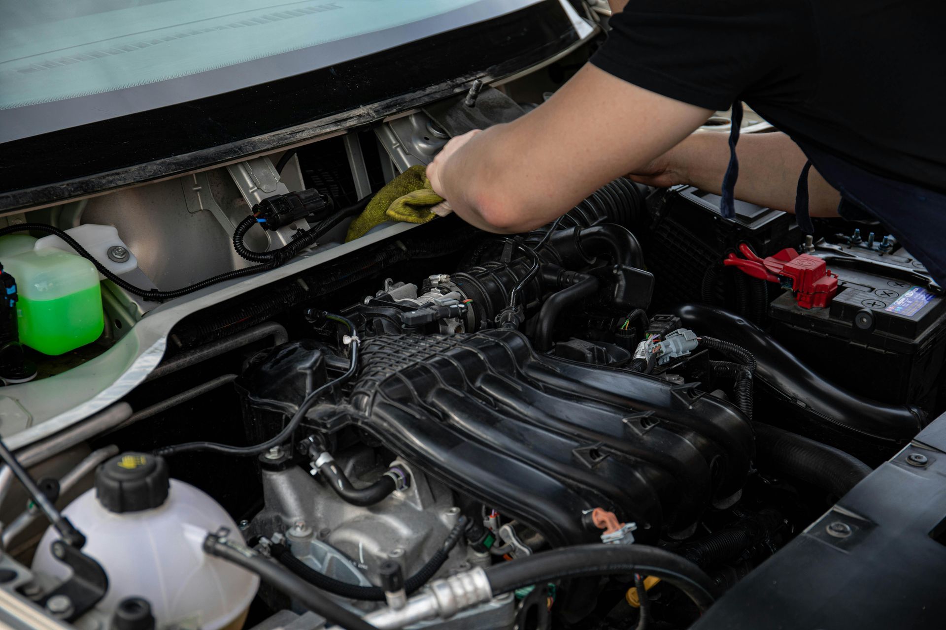 Person cleaning a car engine bay; black engine components, green liquid container, battery visible.