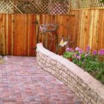 A brick walkway with a wooden fence and flowers in the background.