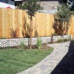 A wooden fence surrounds a lush green yard with a stone wall.