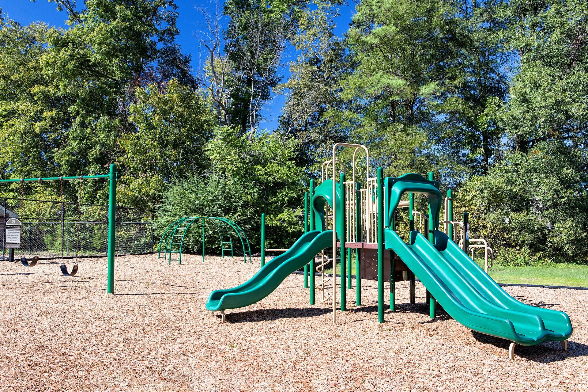 Playground with green slides, swings, and climbing structures on wood chips, surrounded by trees.