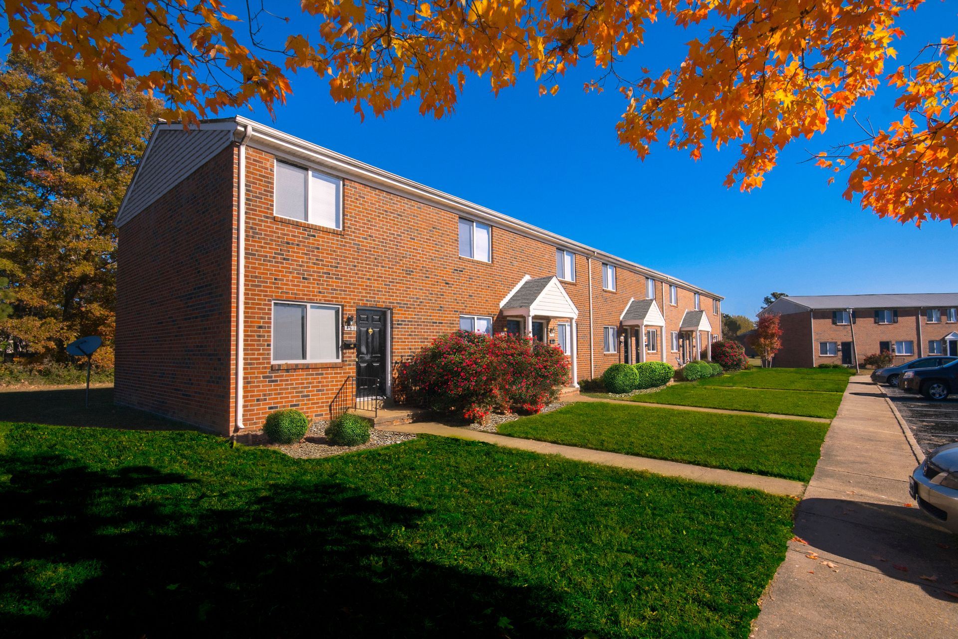 Brick townhouses with green lawns, fall foliage, and a blue sky.