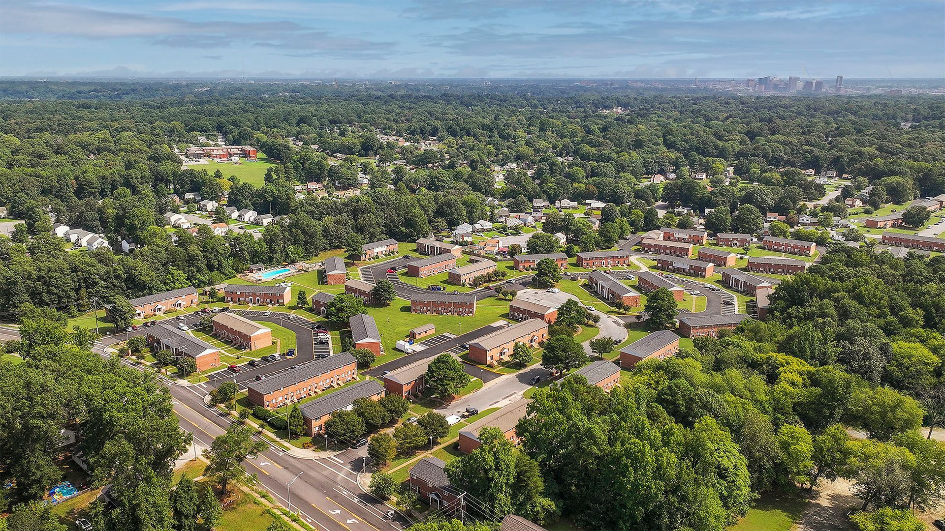 Aerial view of a suburban neighborhood with brick buildings surrounded by green trees and a visible skyline in the distance.