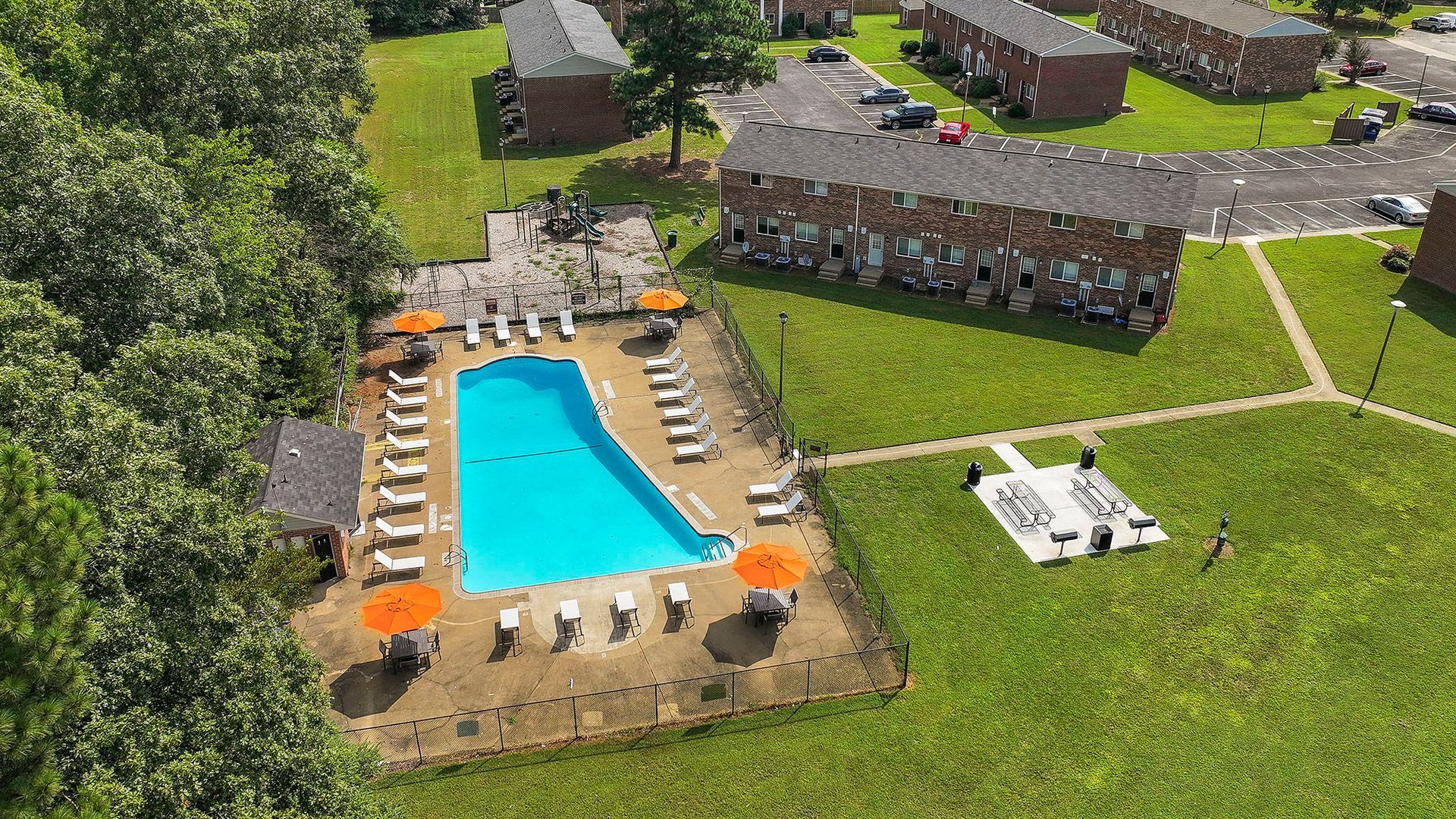 Aerial view of a swimming pool with lounge chairs and orange umbrellas, surrounded by grass and apartment buildings.