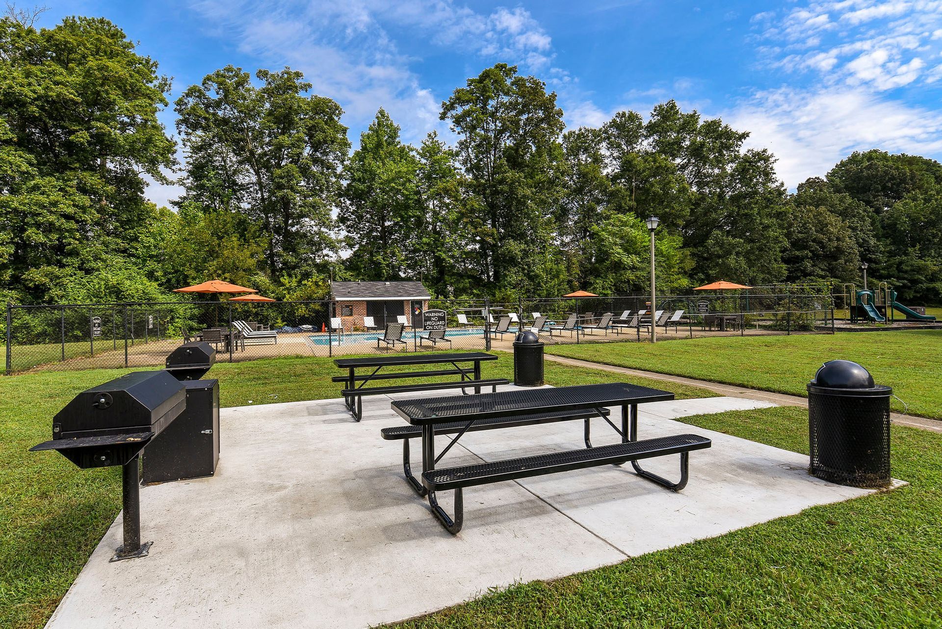 Picnic area with grill, tables, trash can, and fenced pool area in background.