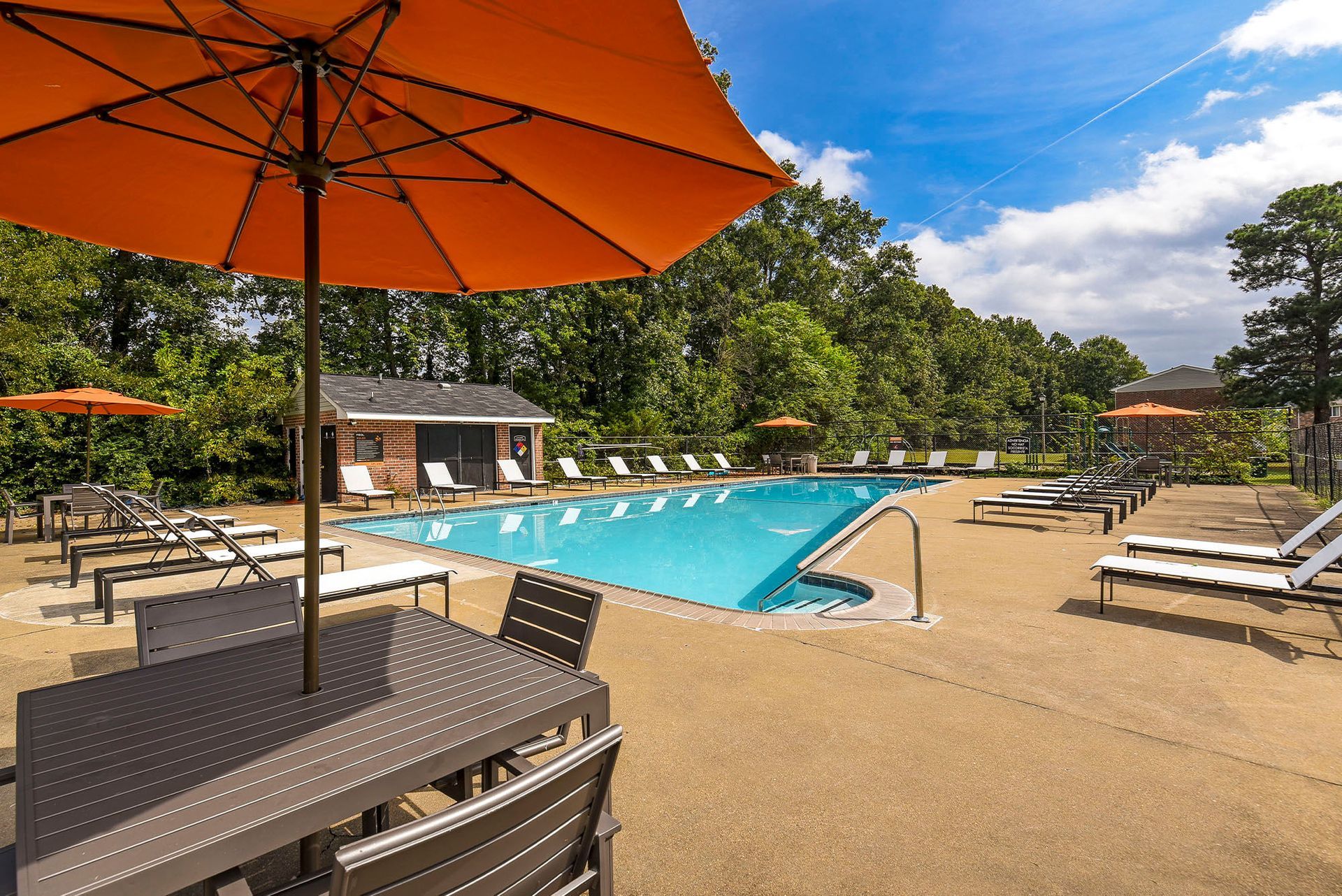 Swimming pool area with orange umbrellas, lounge chairs, and a small building under a blue sky.