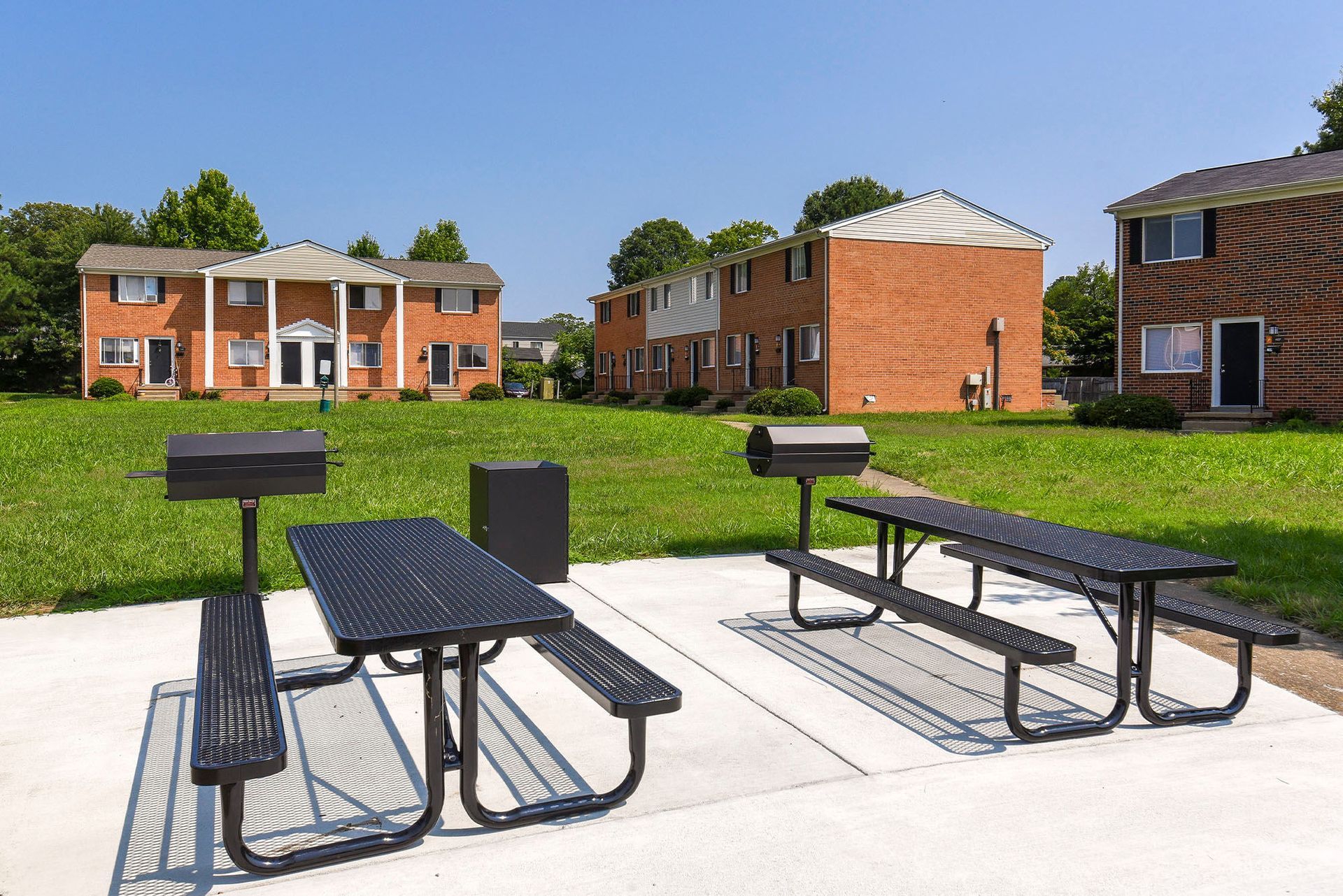 Picnic tables and grills on a concrete pad, with brick apartment buildings and grassy lawn in the background.