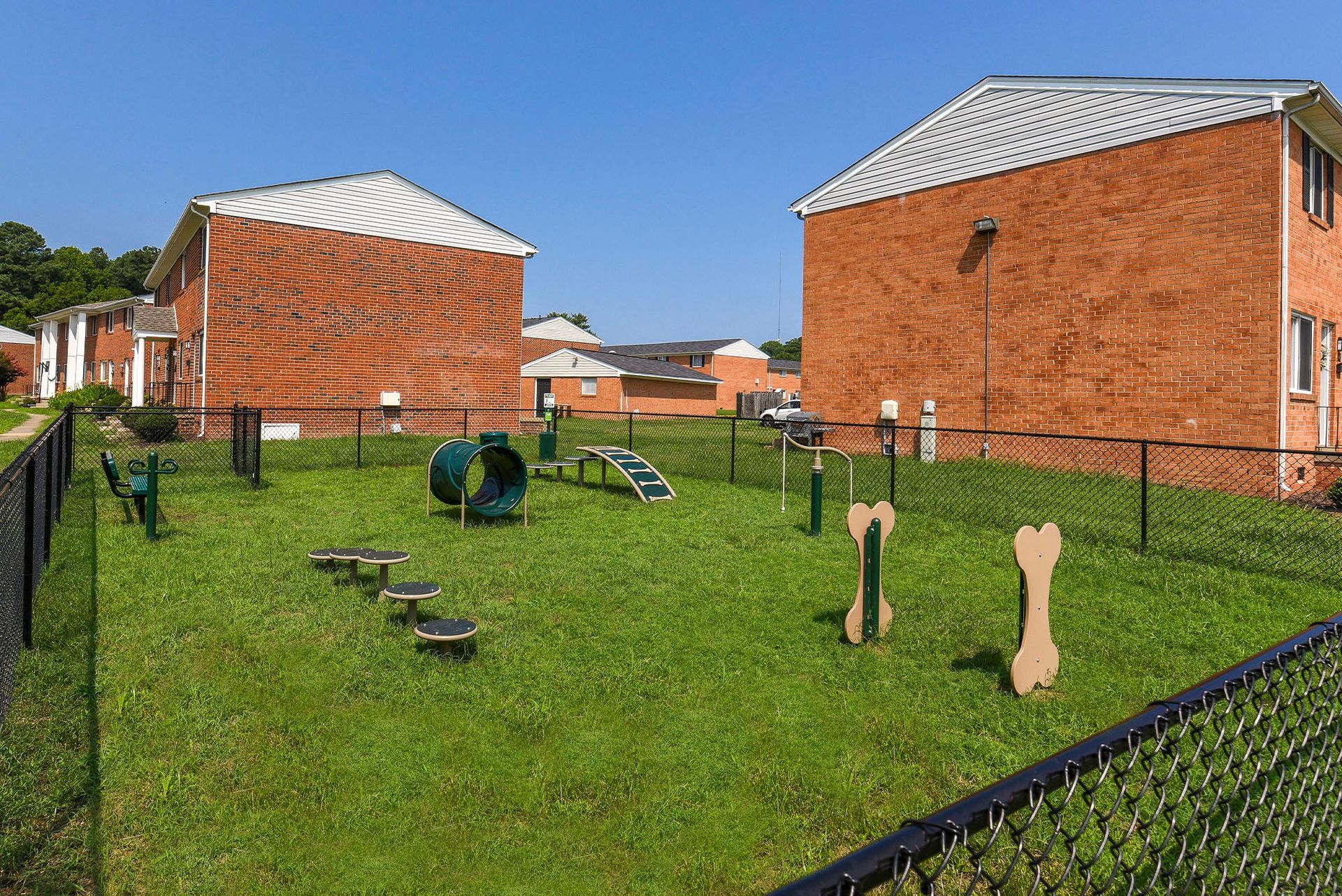 Dog park with agility equipment, green grass, and brick apartment buildings.
