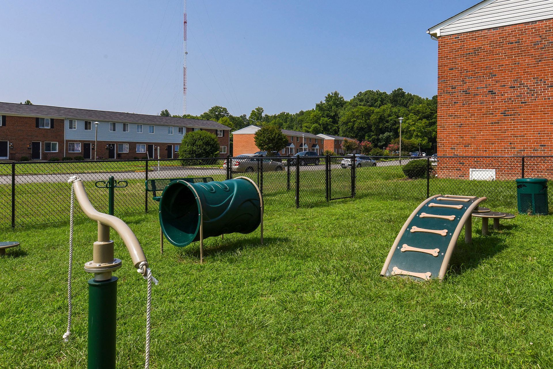 Dog park with agility equipment on grass, fenced in, with apartments in the background.