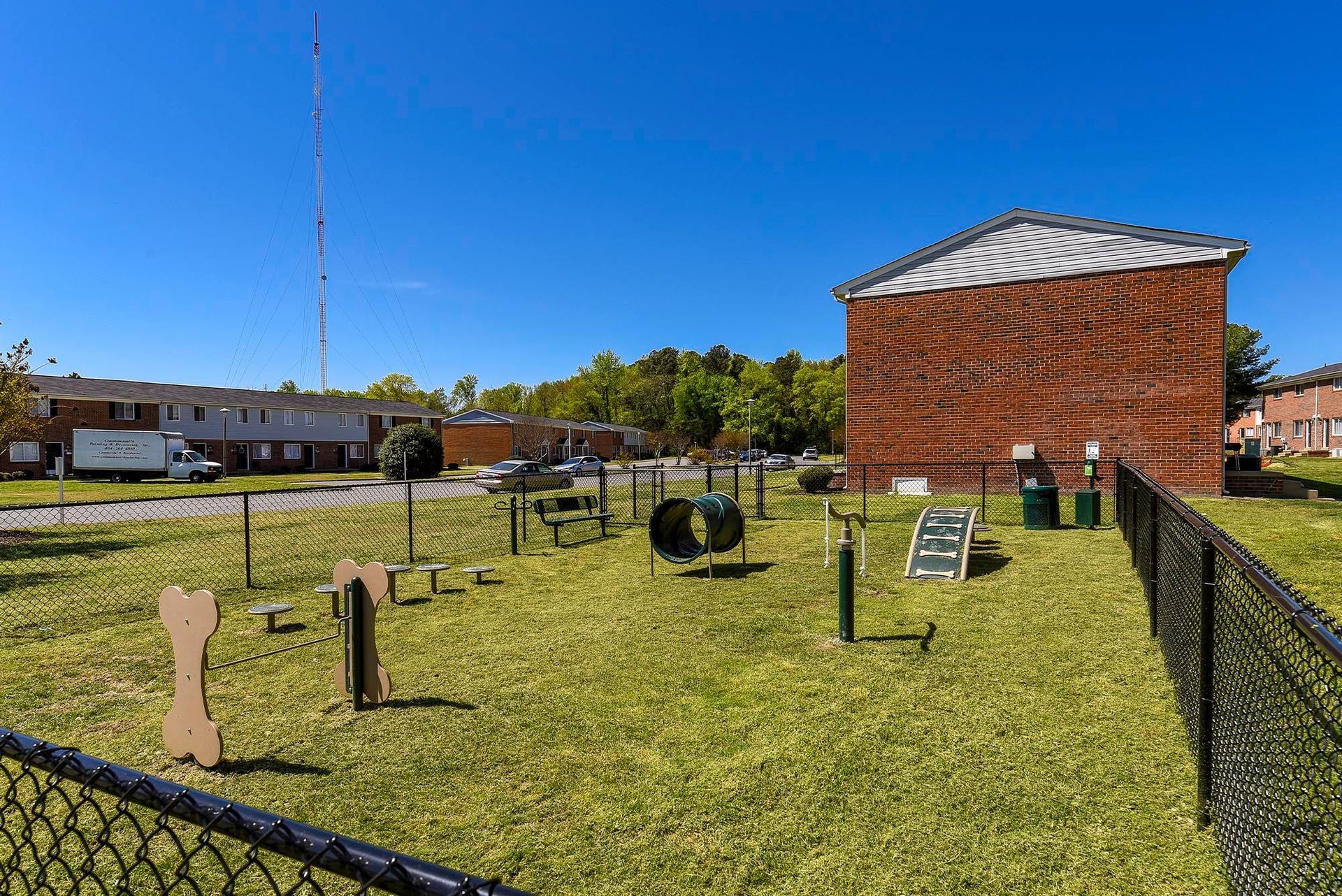 Dog park with agility equipment on green grass, enclosed by a black fence, with brick building backdrop under a blue sky.