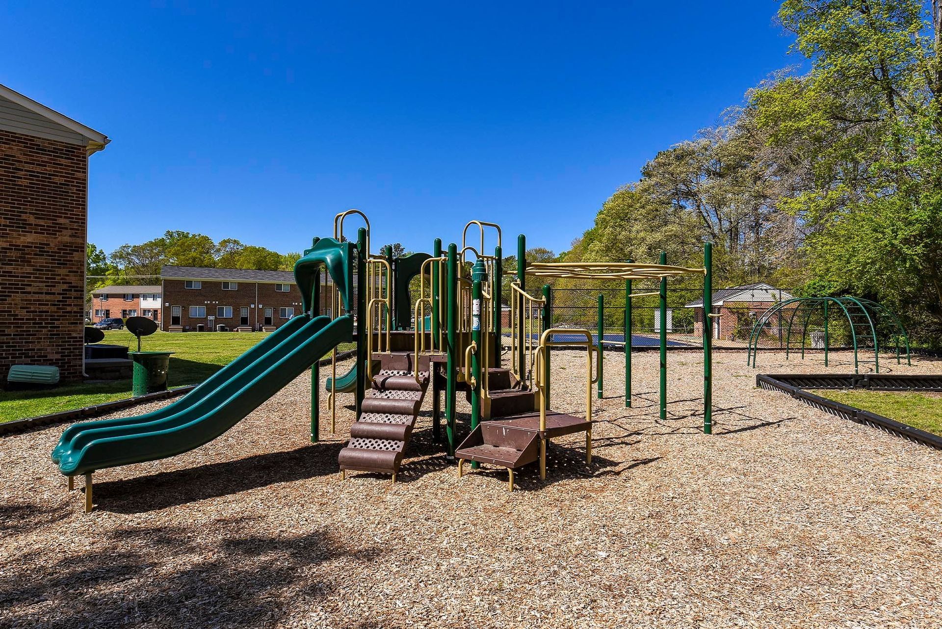 Playground with green slide, stairs, and climbing structure on wood chips, under blue sky.