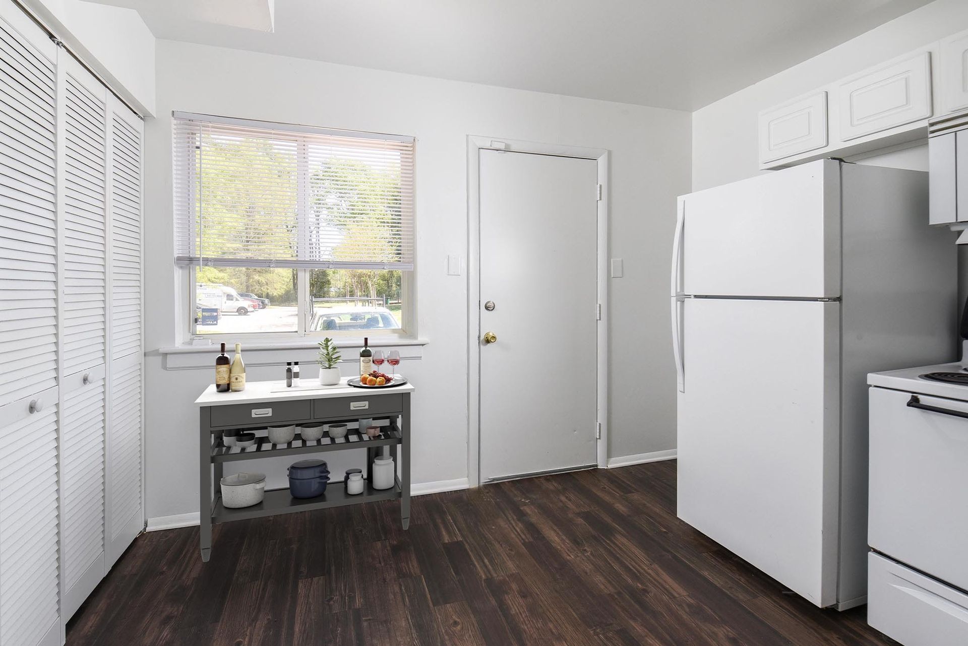 White kitchen with dark wood-look flooring. White cabinets, refrigerator, and door. Window with sheer curtain. Gray serving cart.