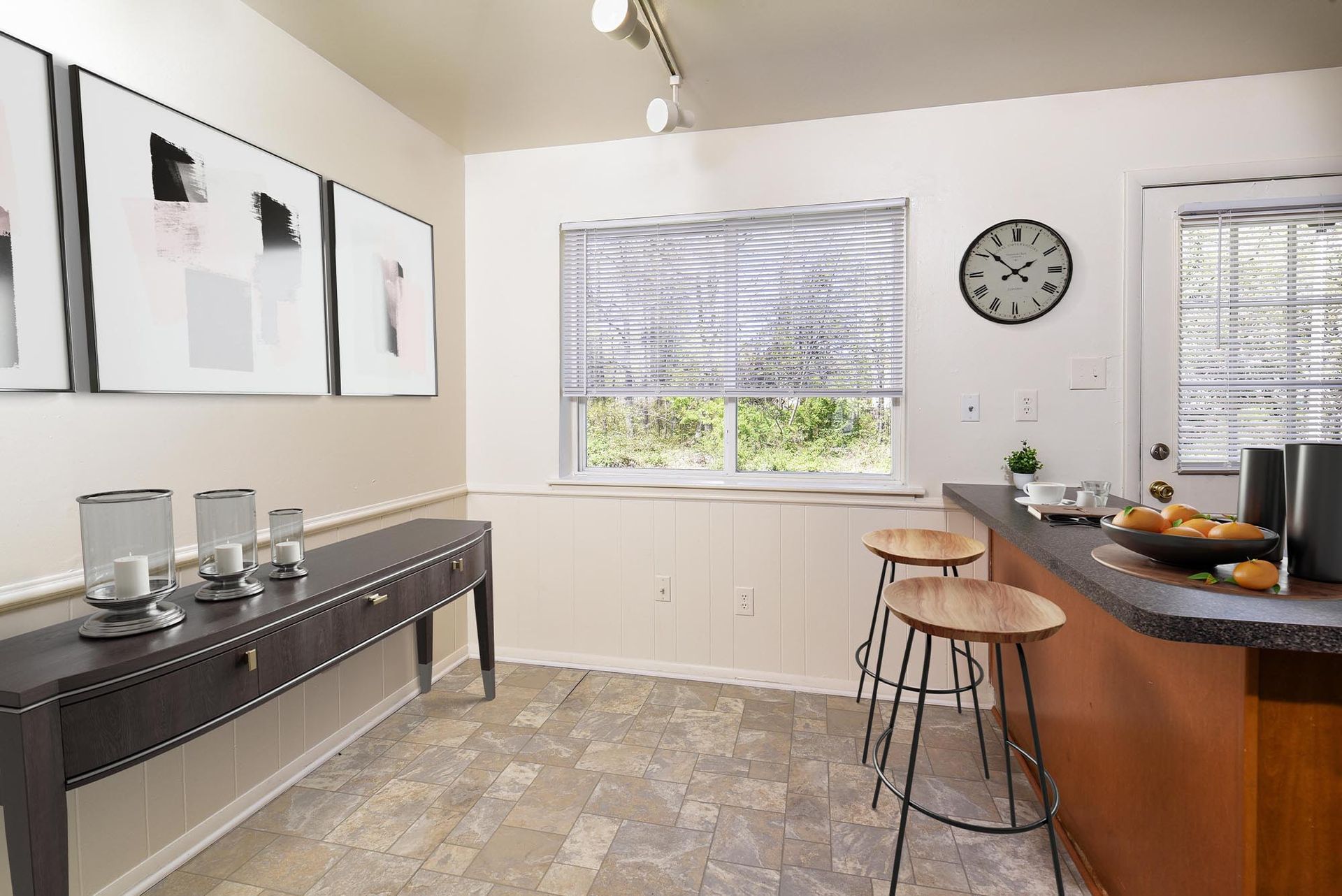 Kitchen with a bar, stools, and a table with candles. Artwork on the wall and a window with a blind.