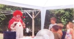 A clown is standing in front of a group of children under a white tent.