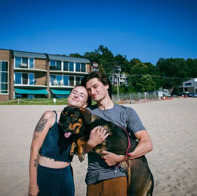 Couple on beach, holding dog, posing with building backdrop. Clear blue sky, sunny.
