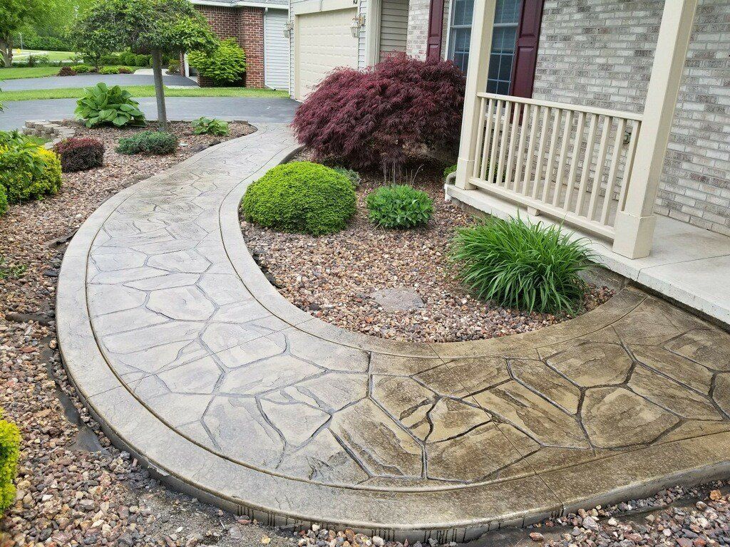 Curved stone walkway leading to a house with landscaping; gray concrete with a stone pattern.