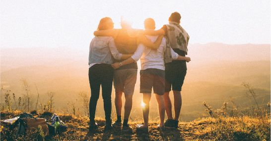 Four people with arms around each other, standing on a hilltop, watching the sunset.