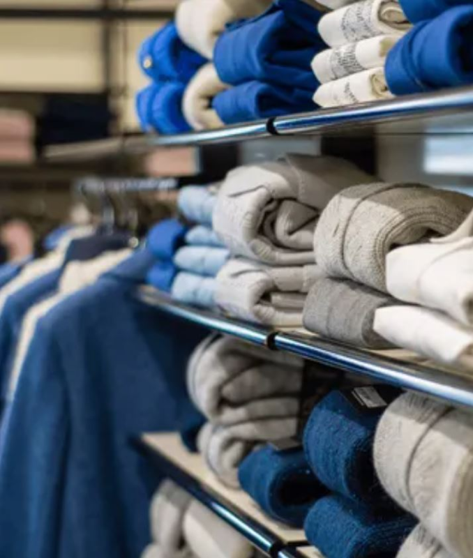 Scaffali pieni di maglioni blu, grigi e bianchi piegati in un negozio al dettaglio. Giacche appese su uno scaffale lì vicino.