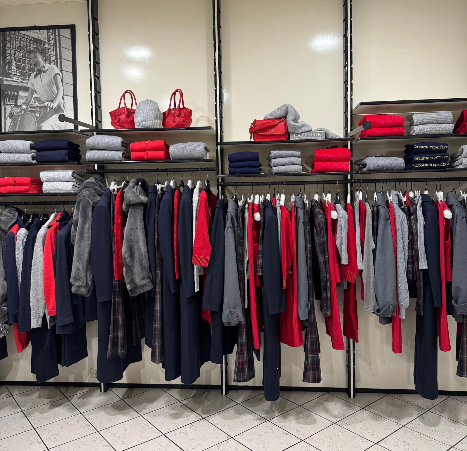 Espositore per negozio di abbigliamento: scaffali con abiti rossi, grigi e blu navy. Scaffali con abiti piegati e borse.