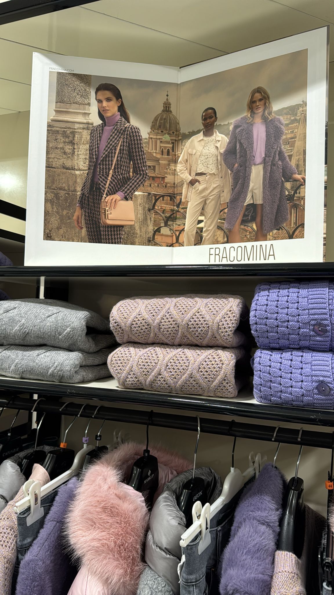 Esposizione di abiti: maglioni sugli scaffali, cappotti sulle grucce, pubblicità che mostra modelle di fronte a un edificio.