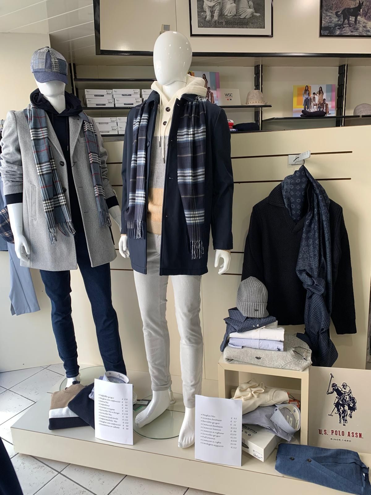 Esposizione di un negozio di abbigliamento: manichini in abiti invernali, tra cui cappotti, sciarpe, cappelli e maglioni, con listini prezzi.