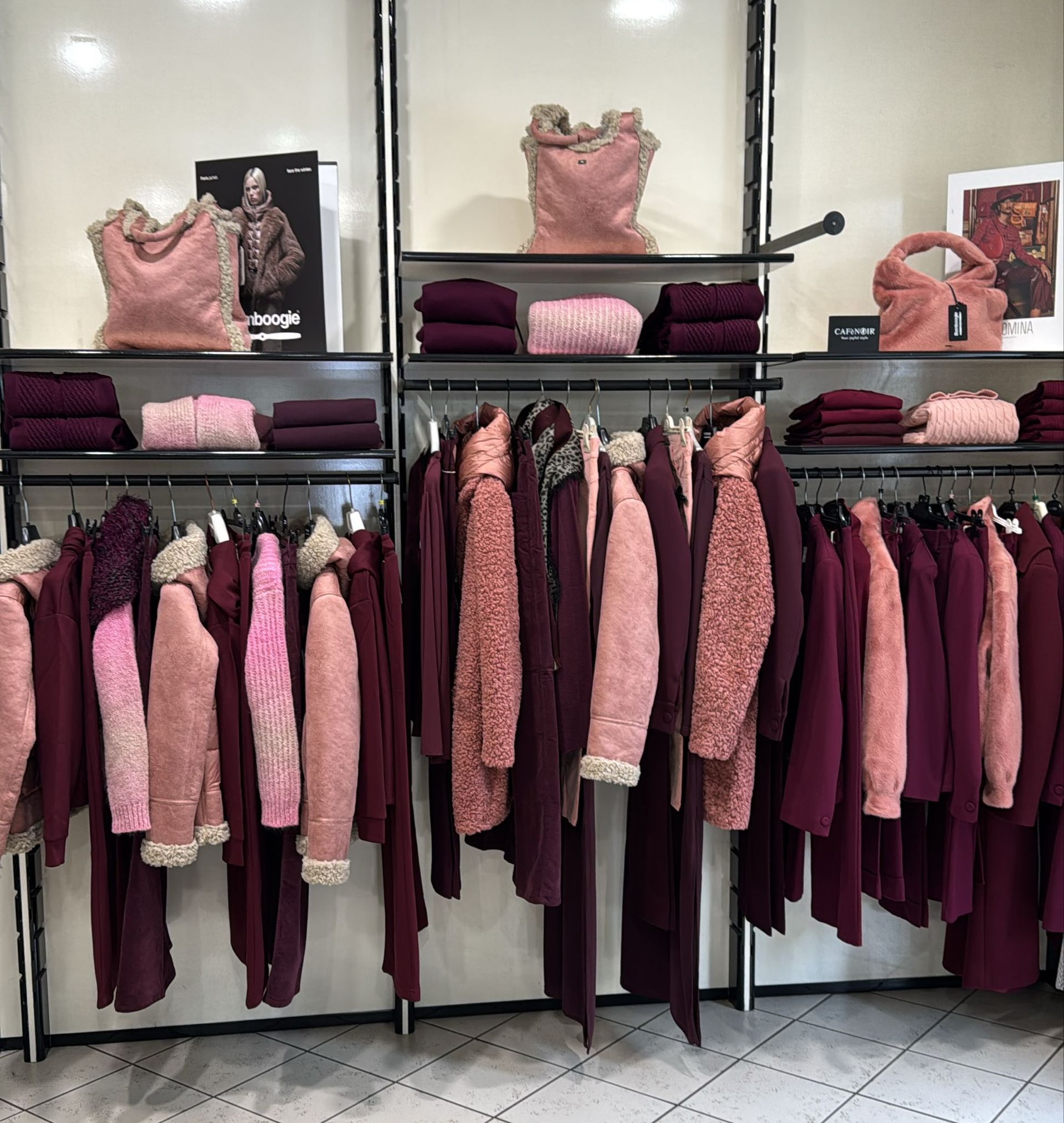 Esposizione di un negozio di abbigliamento con scaffali pieni di cappotti e scaffali pieni di maglioni piegati, tutti nei toni del rosa e del bordeaux.