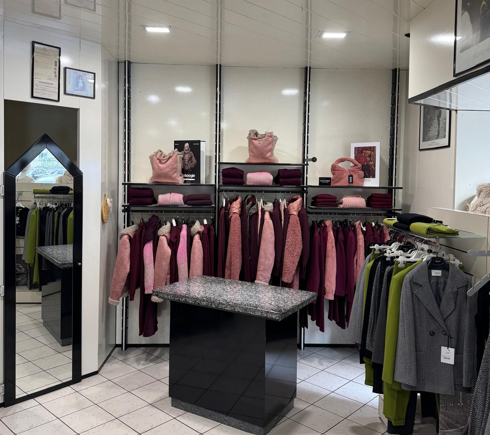 Interno di un negozio di abbigliamento con scaffali pieni di abiti rosa e bordeaux e scaffali espositivi con borse.