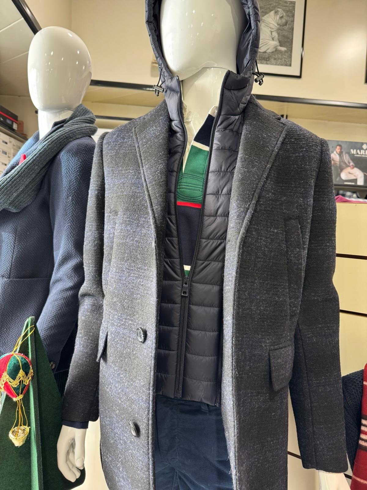 Manichino in un negozio che indossa un cappotto grigio sopra un gilet nero e un maglione con cappuccio; un altro manichino è sulla sinistra.