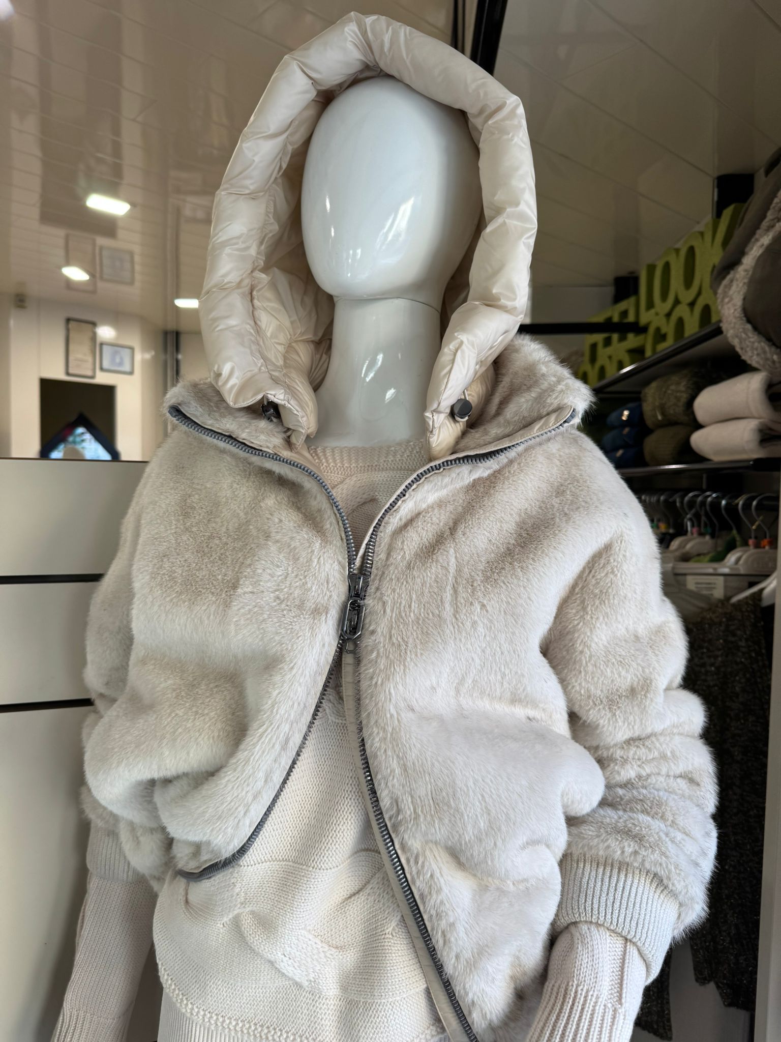 Manichino con una giacca soffice color crema con cerniera e cappuccio, all'interno di un negozio di abbigliamento.