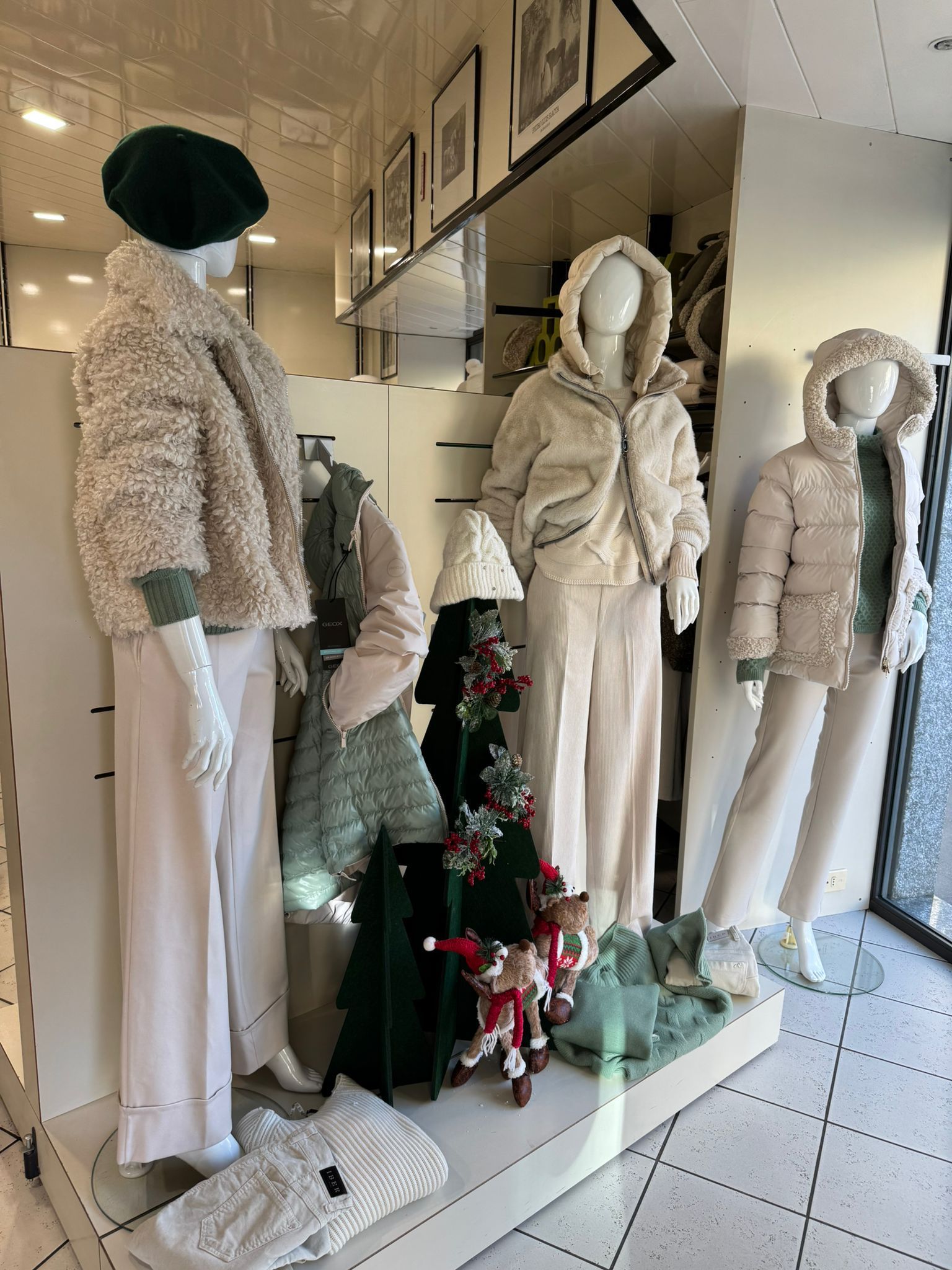 Vetrina di un negozio di abbigliamento con manichini che indossano capispalla e pantaloni chiari. Davanti c'è un piccolo albero di Natale.