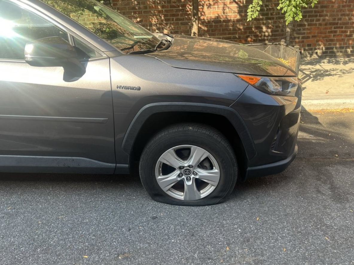 Man changing a car tire on a wet road, woman holding an umbrella nearby.