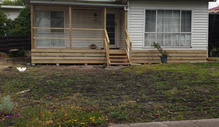 Wooden porch with steps leading to a white house with horizontal siding, and a patchy lawn in front.