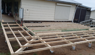Deck construction in progress; wooden frame on adjustable footings, attached to a house with white siding.