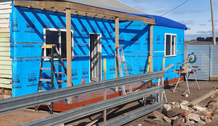 House under construction, blue protective wrap, wooden beams, ladder, sunny outdoor setting.