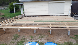 Wooden deck frame construction in front of a white building, set on concrete pillars.