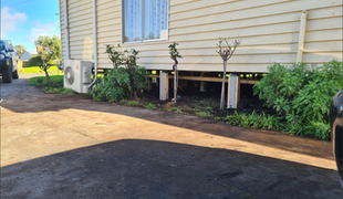 Side view of a light beige house with an air conditioning unit and some plants growing below the window.