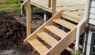 Wooden stairs leading up to a deck attached to a house; surrounded by dirt and grass.