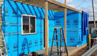 Exterior of building under construction; blue protective wrap, window, wood frame, ladder.