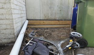 Tools and debris in a dirt area beside a brick wall, wooden beam, and a green trash bin.
