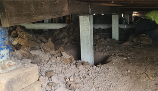 Dirt and debris under a house with support beams. A person in a green shirt is visible.