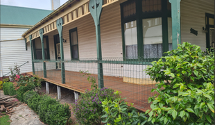 A wooden house with a porch. The house is painted white and green, with a brown wooden deck and green bushes.