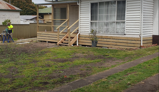 House with wooden porch and steps; person working in yard; grassy lawn and sidewalk.