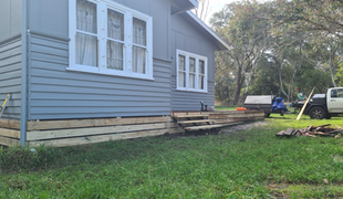 Gray house with new wooden base, steps, and windows. Lawn in front. Truck and trailer in background.