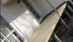 Side view of a white house with a partially constructed deck. Sunlight casts shadows on the house and deck.