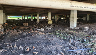 Underneath a house: dirt ground, concrete pillars, wooden beams, PVC pipe, and debris.