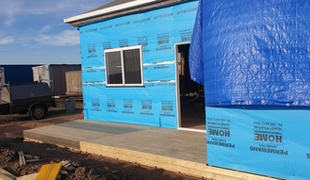 Building exterior under construction with blue protective wrap, concrete patio, and a covered doorway.