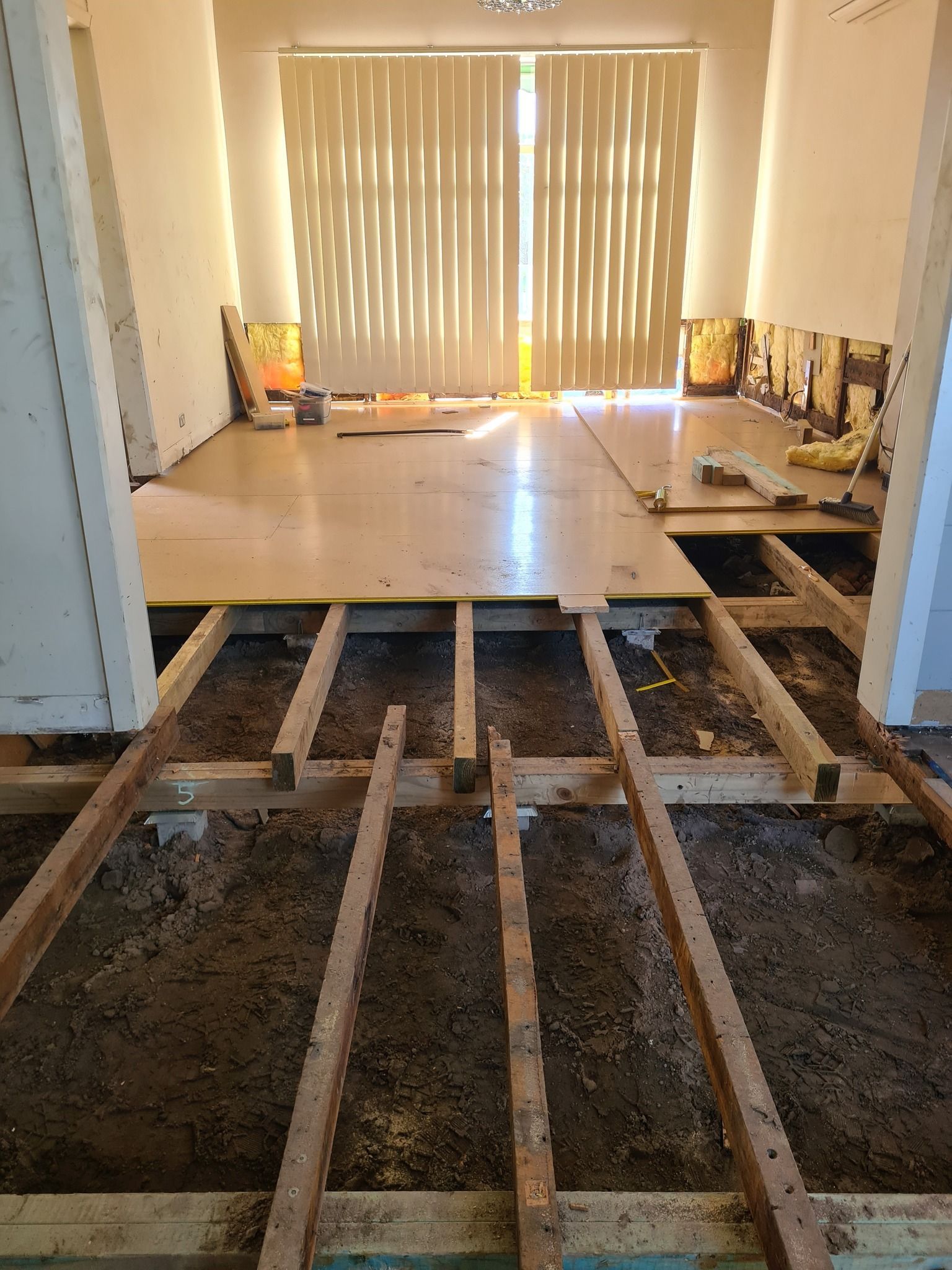Room under construction, with exposed floor joists, plywood subfloor, and a doorway with vertical blinds.