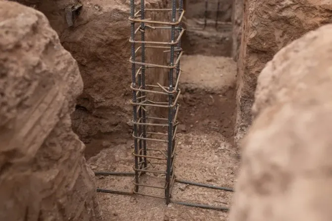 A steel rebar cage for a vertical column sits inside a narrow, dirt-walled trench prepared for a concrete foundation.